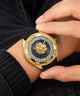 A close-up of a wrist wearing a luxury watch with a gold-tone case, a black strap, a black sunburst dial, a gold lion emblem, and crystal accents.