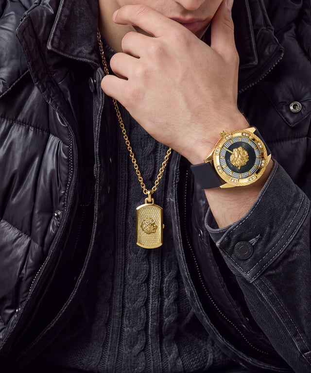 A close-up lifestyle shot of a person wearing a gold-tone watch with a black dial and bezel, along with a matching gold dog tag necklace. The person is wearing a black puffer jacket and a black knit sweater.