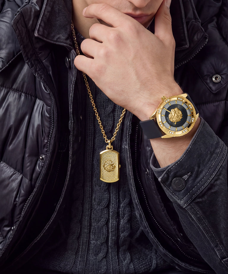 A close-up lifestyle shot of a person wearing a gold-tone watch with a black dial and bezel, along with a matching gold dog tag necklace. The person is wearing a black puffer jacket and a black knit sweater.