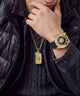 A close-up lifestyle shot of a person wearing a gold-tone watch with a black dial and bezel, along with a matching gold dog tag necklace. The person is wearing a black puffer jacket and a black knit sweater.