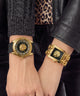 Two gold-tone fashion watches with black faces and lion-head motifs worn on a person's wrists, styled with a black leather jacket, leopard-print top, and gray knit sweater.