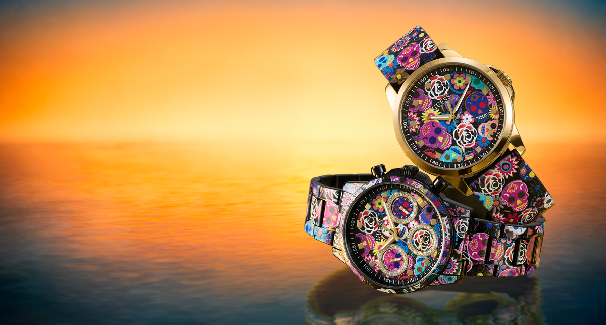 Two ornate wristwatches with colorful floral and mechanical dial designs and patterned straps, displayed on a reflective surface against a warm orange sunset background.